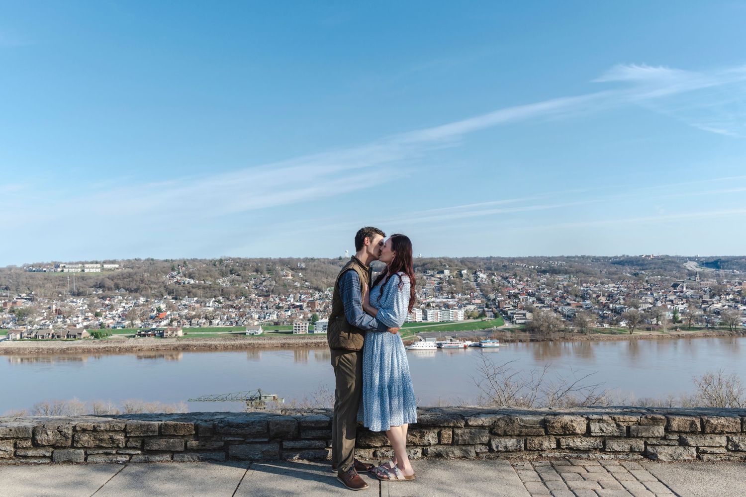 Couple sharing a kiss on a scenic overlook with a river and city skyline under a blue sky.
