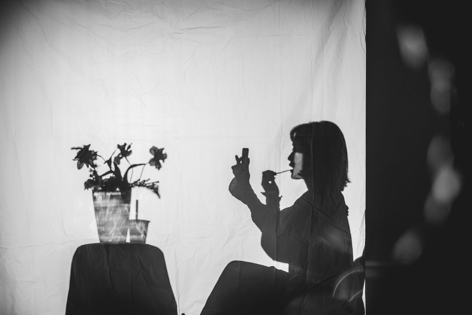 Black and white photo of a woman taking a selfie beside a potted plant against a white backdrop.