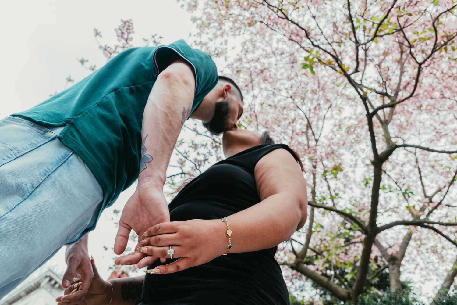 A low-angle view of two people holding hands beneath a blooming pink cherry blossom tree outdoors.