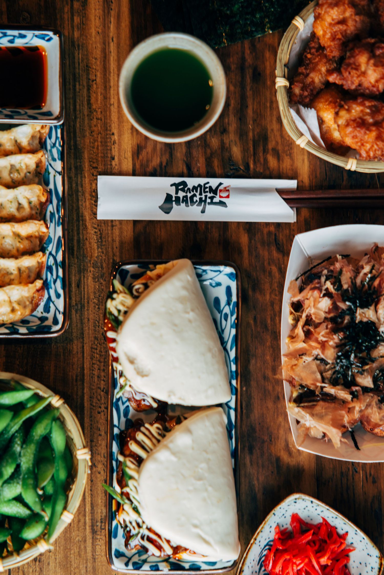 Overhead view of Japanese bao buns, spring rolls, edamame, and fried chicken on a wooden restaurant table.