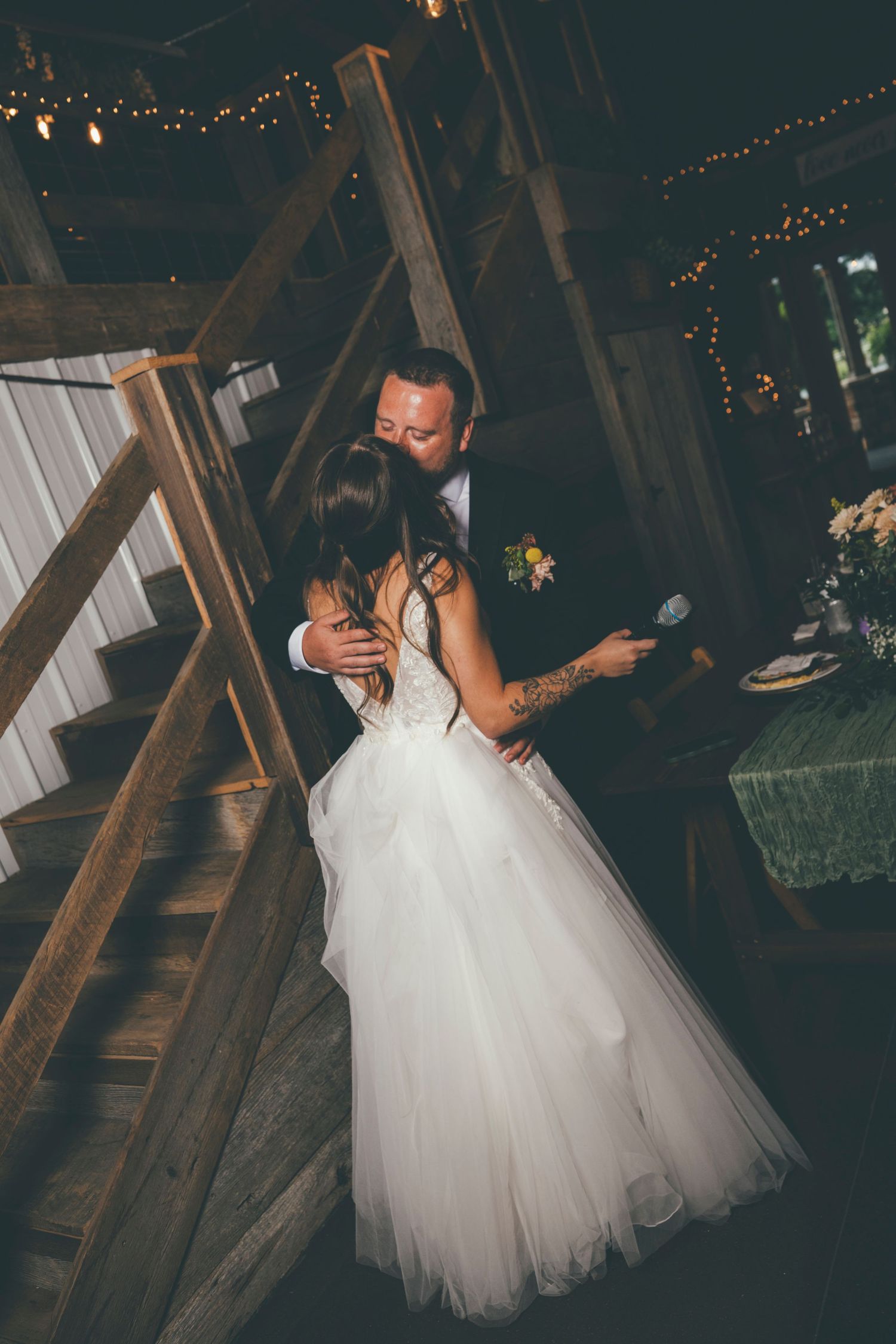 Bride and groom share romantic first dance in rustic barn wedding venue with string lights and wooden staircase.