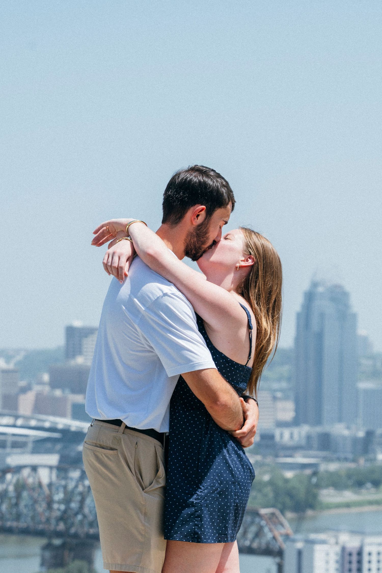 Couple sharing a romantic kiss with Cincinnati skyline and river bridge in the background on a sunny day.