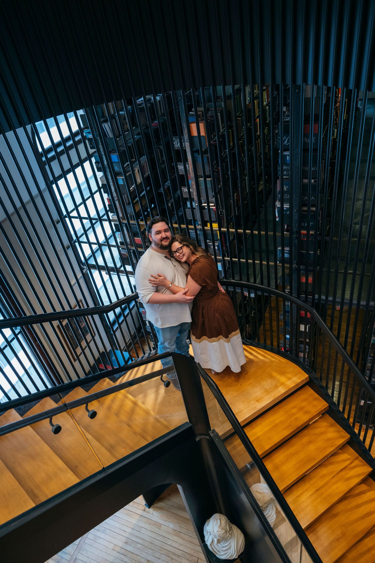 Couple embracing on a wooden spiral staircase, smiling at camera from above in a modern building.