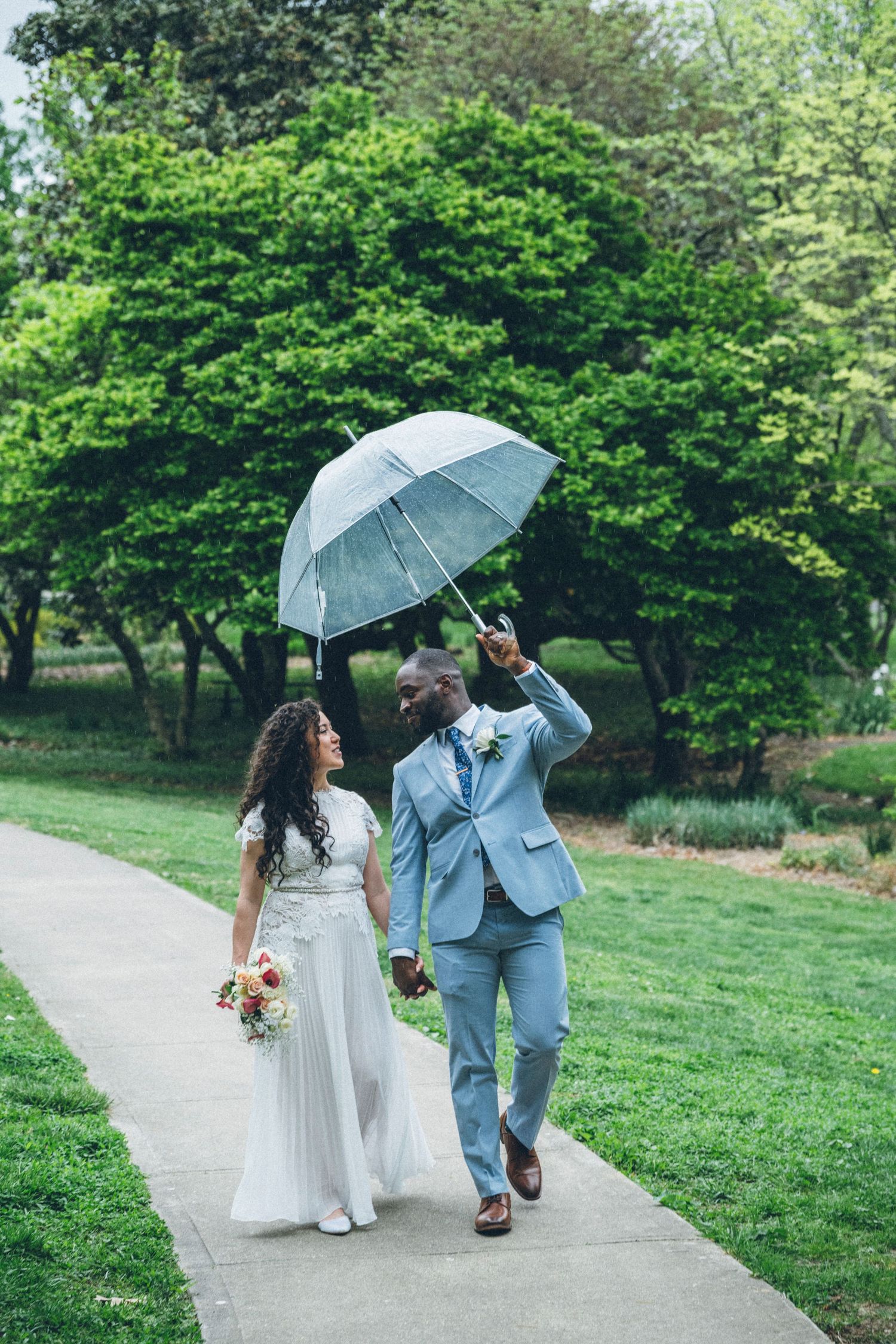 Happy interracial newlyweds walk hand-in-hand under a clear umbrella on a garden path on their wedding day.