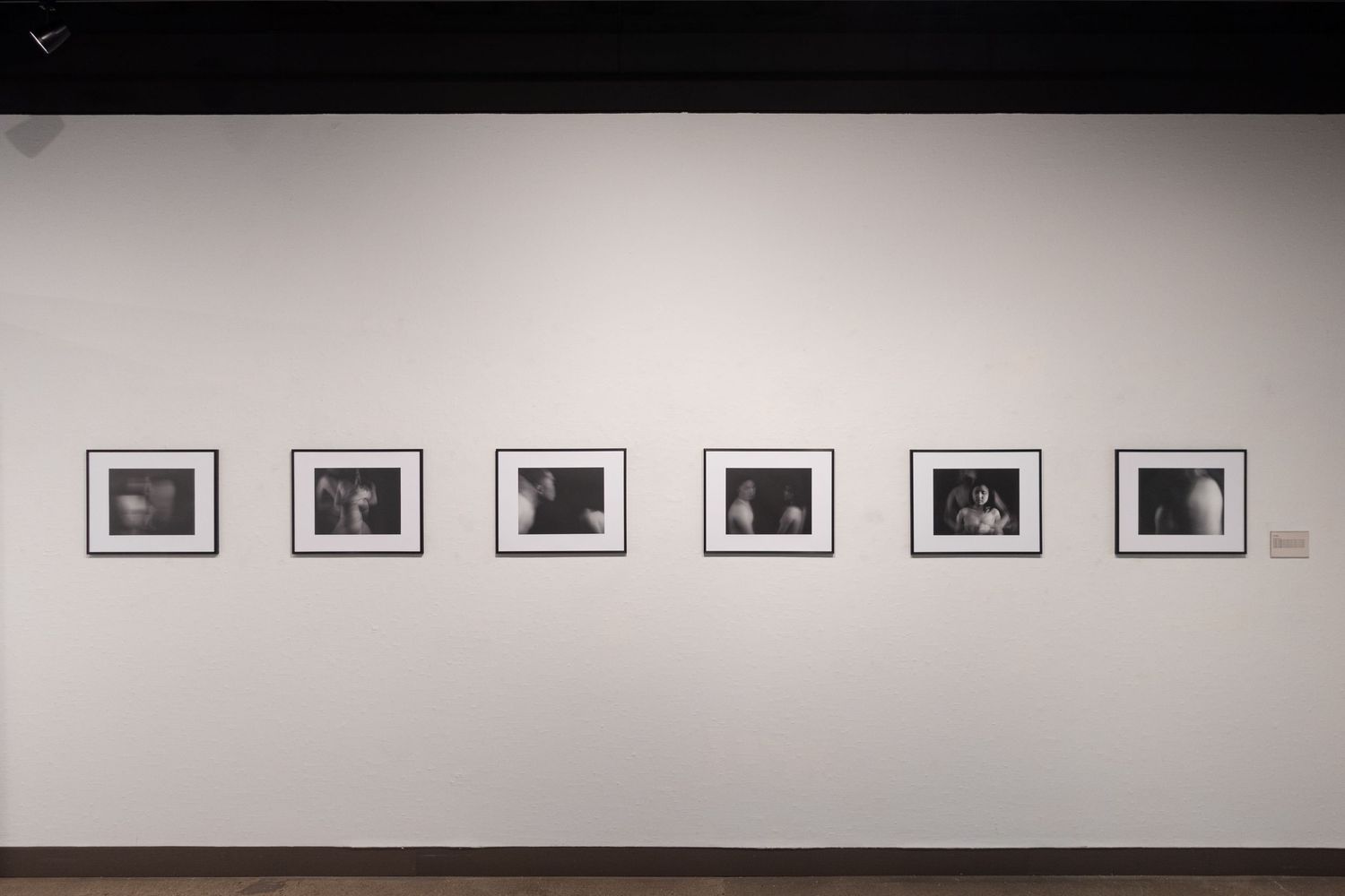 Six black-and-white framed photographs displayed in a row on a white gallery wall under dim lighting.