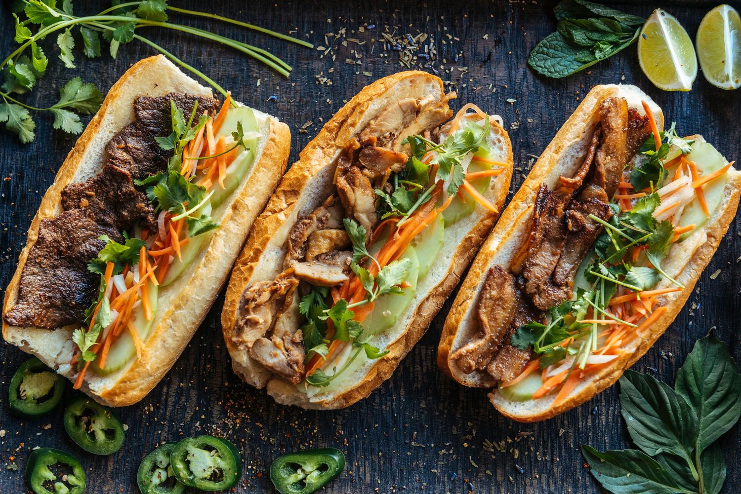 Three banh mi sandwiches topped with grilled meat, pickled carrots, fresh herbs, and jalapenos on a dark wooden surface.