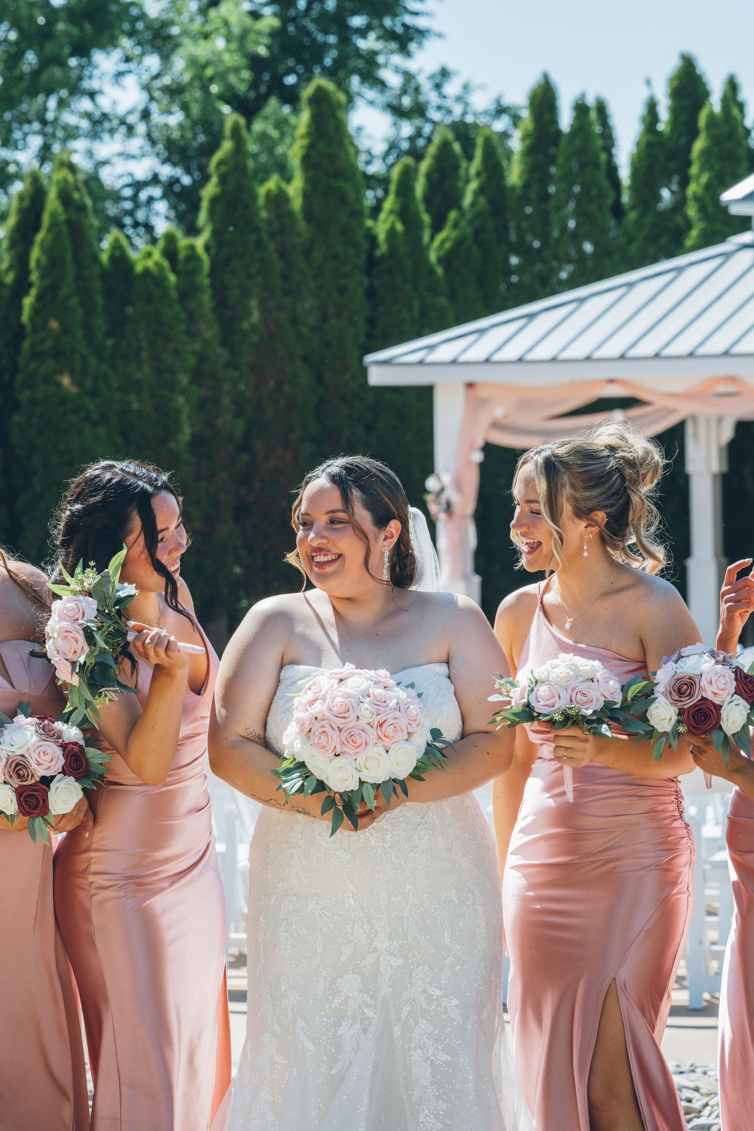 Bride in white lace gown holds pink bouquet, laughing with bridesmaids in blush satin dresses outdoors.