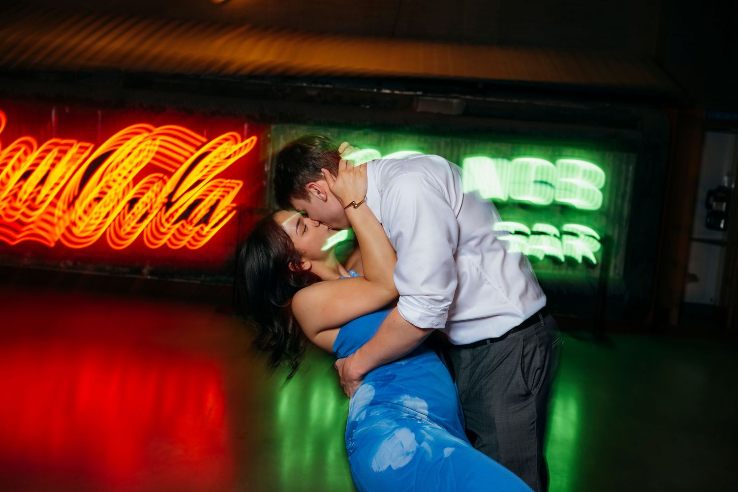 Couple sharing a romantic kiss in front of glowing Coca-Cola neon sign at a dimly lit venue.