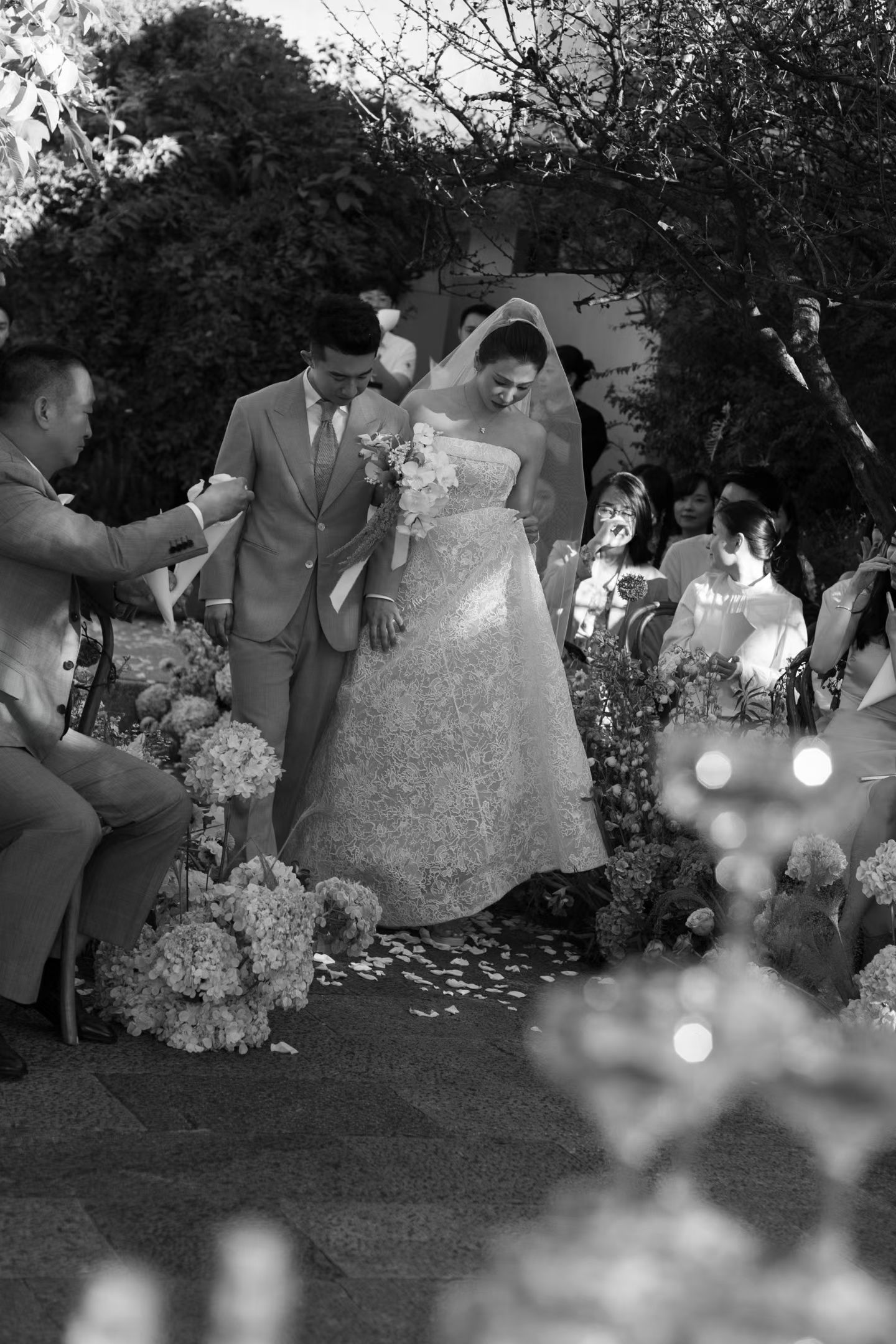 Black and white photo of bride walking down outdoor wedding aisle with escort amid floral decorations.