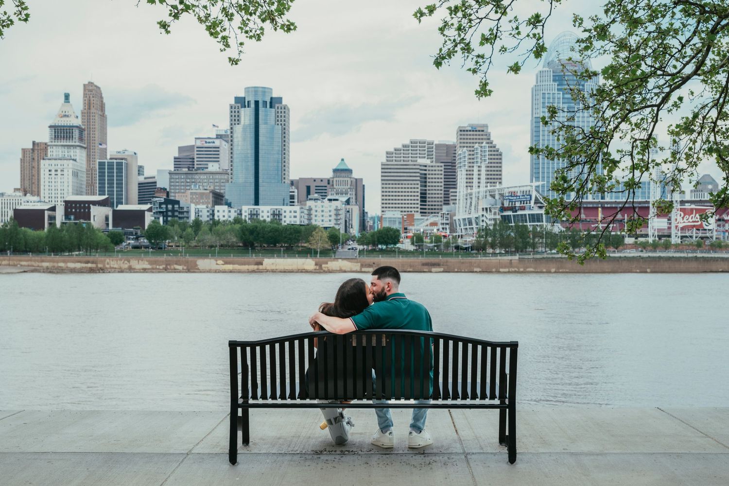Couple sitting closely on a bench by a river with a city skyline in the background.