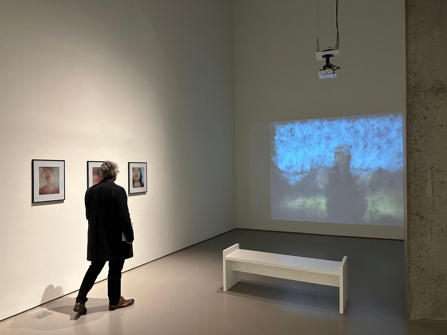 Person viewing framed photos and a blue projected video installation on white gallery walls with a white bench.