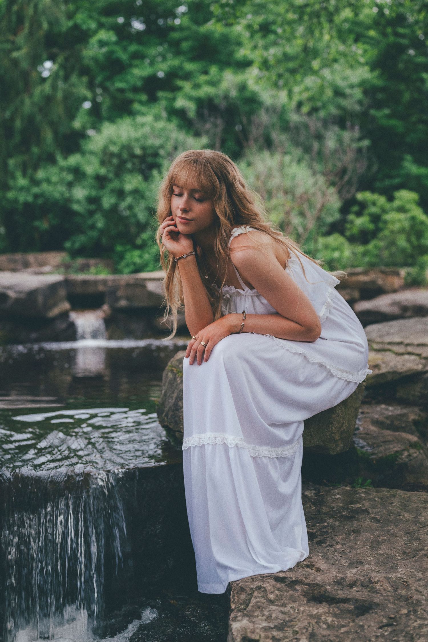 Woman in white dress sitting on rocks by a small waterfall surrounded by lush green trees.