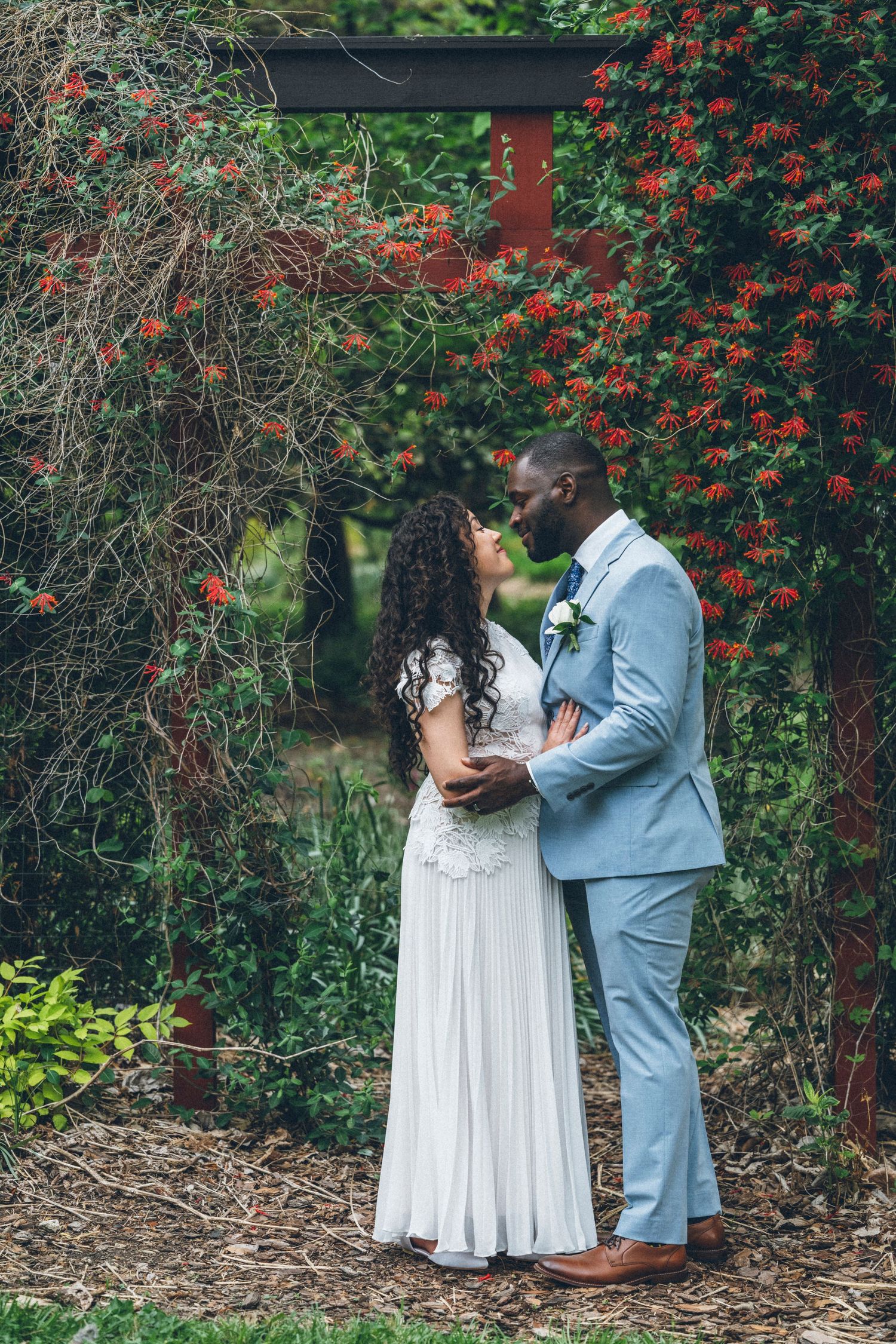 Bride and groom share a romantic moment under a floral arch with red blooms in a lush garden setting.