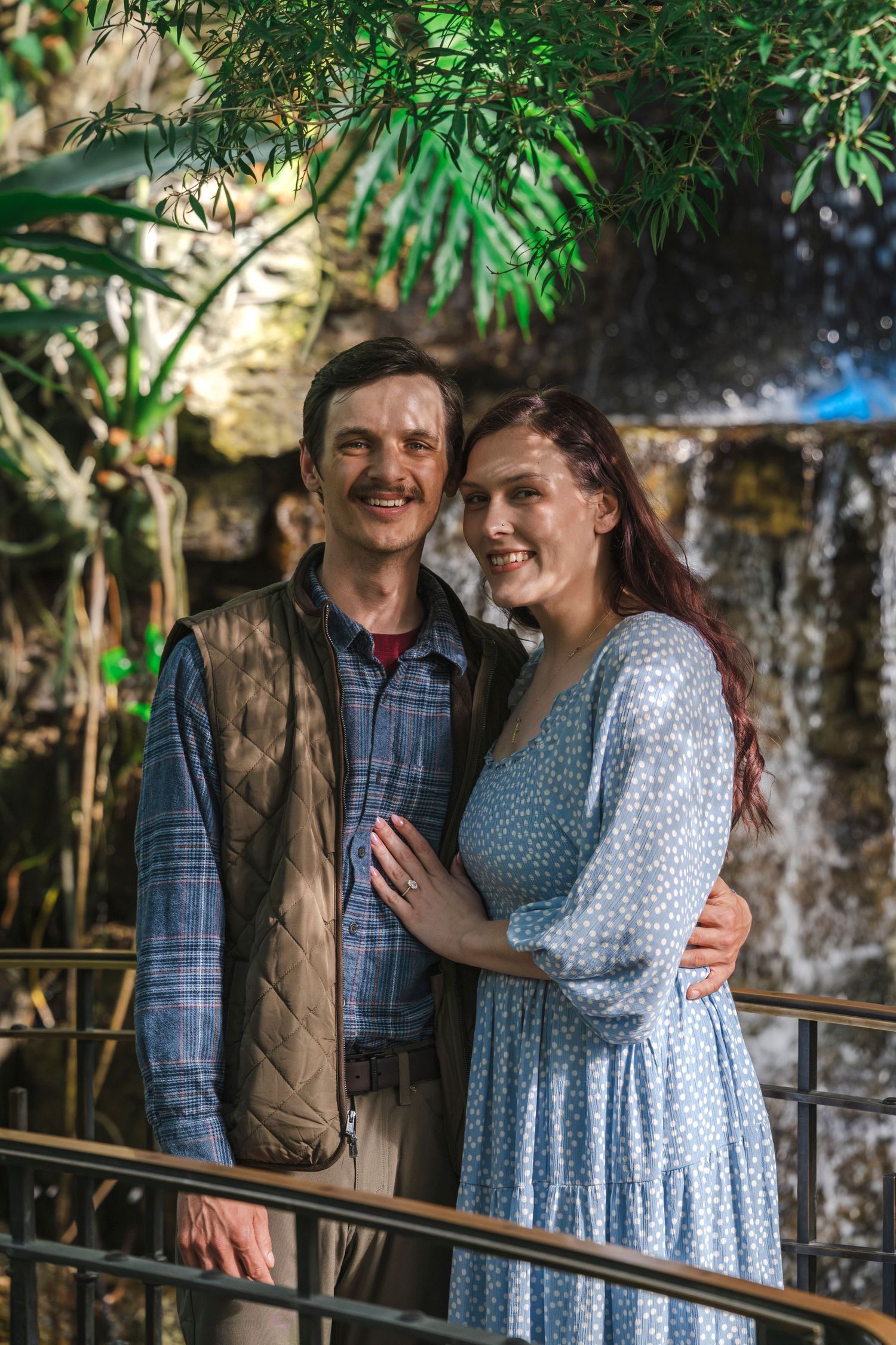 Smiling couple embracing near a waterfall surrounded by lush tropical greenery.