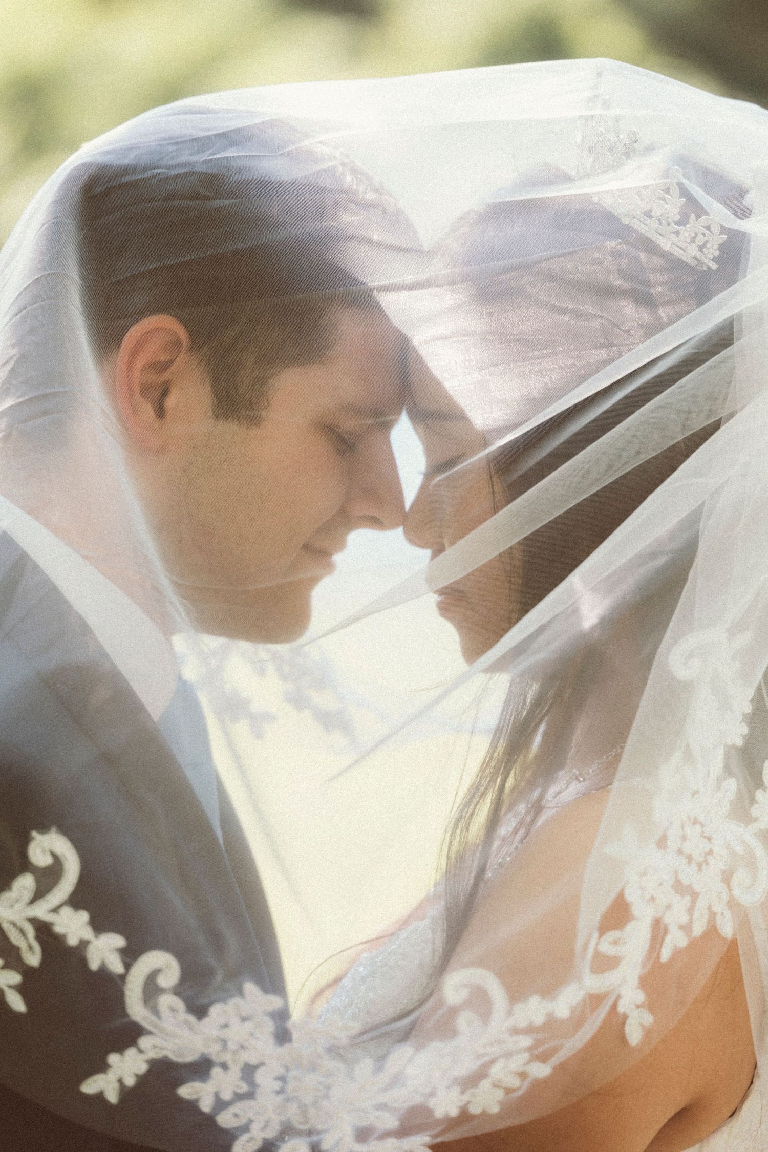 Bride and groom sharing an intimate moment under a lace-trimmed wedding veil, foreheads touching tenderly.