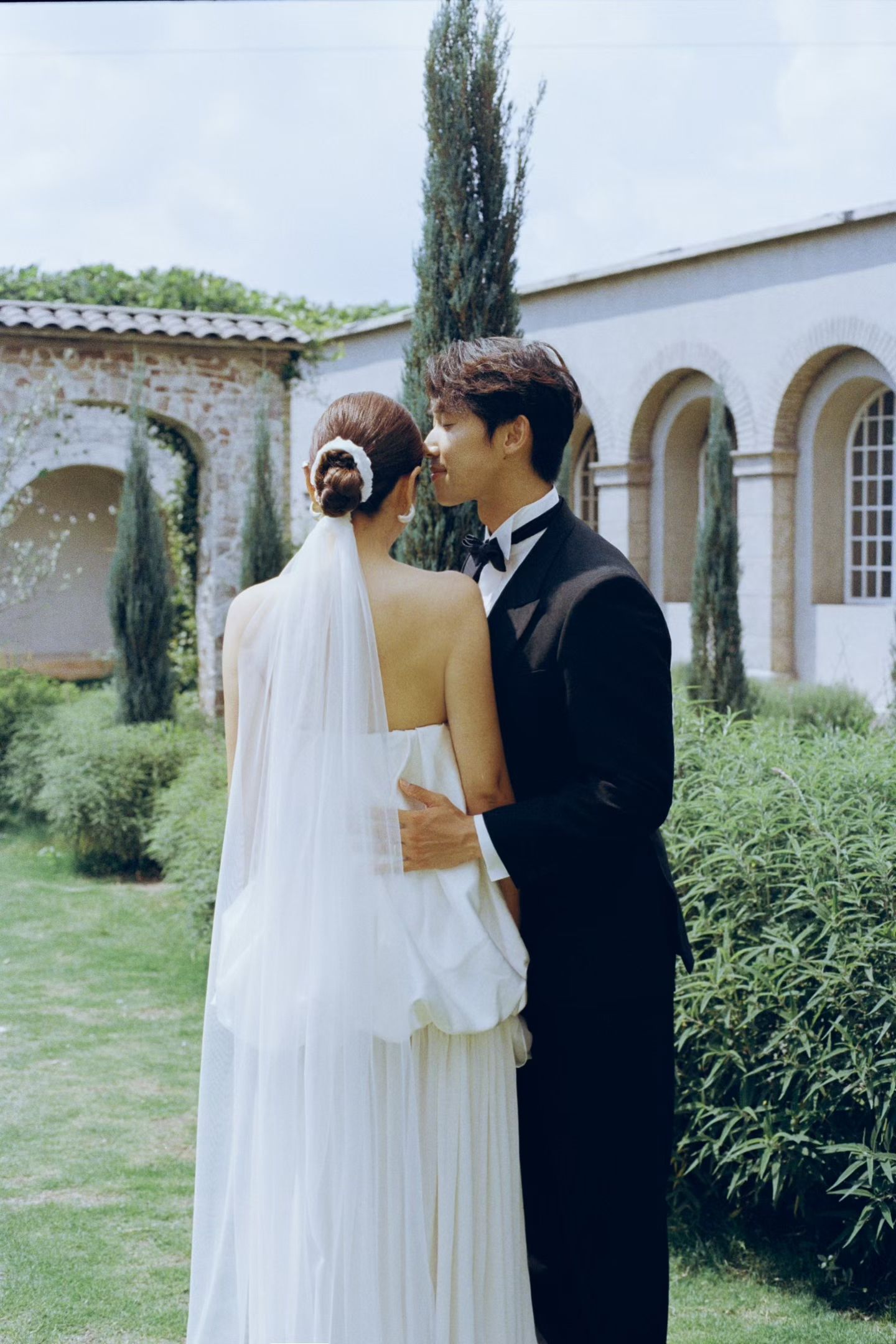 Bride and groom embracing in garden near arched stone building with cypress trees in background.