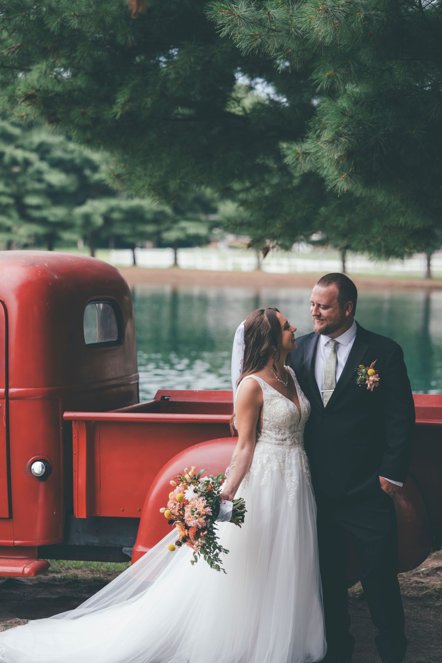 Bride and groom share a romantic moment beside a vintage red truck near a serene pond, bride holding a colorful bouquet.