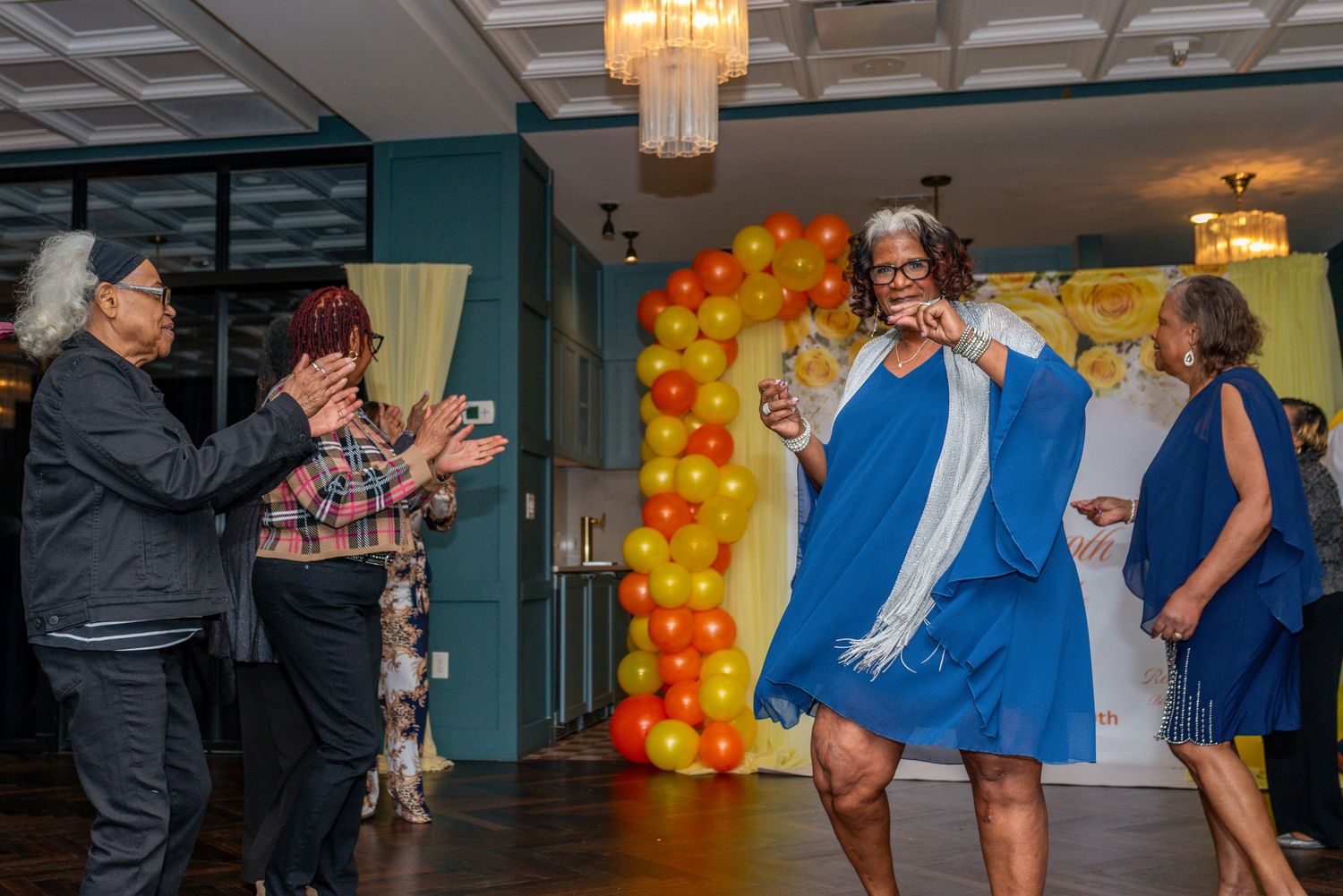 A woman in a blue graduation gown celebrates joyfully at an indoor party with colorful balloon decorations.