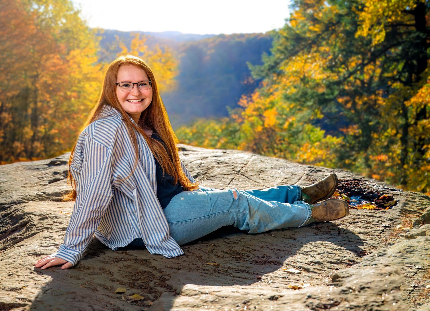 Teen girl with red hair sits on a rock surrounded by colorful autumn foliage during an outdoor fall photo session.