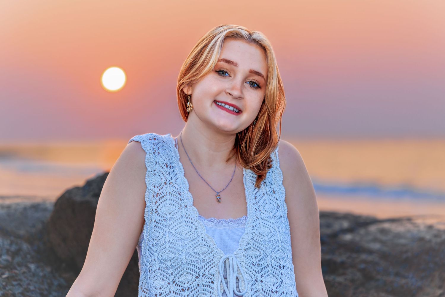 Smiling young woman in white lace top posing on beach rocks during golden sunset.
