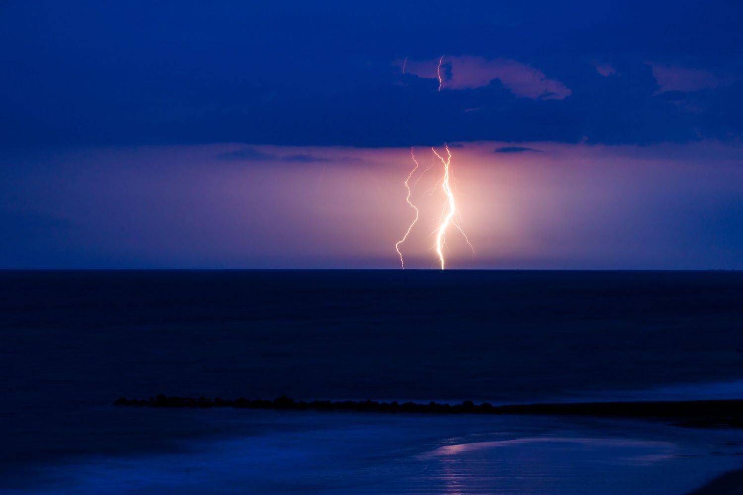 Single powerful lightning bolt striking over the ocean at night with a rocky breakwater below.