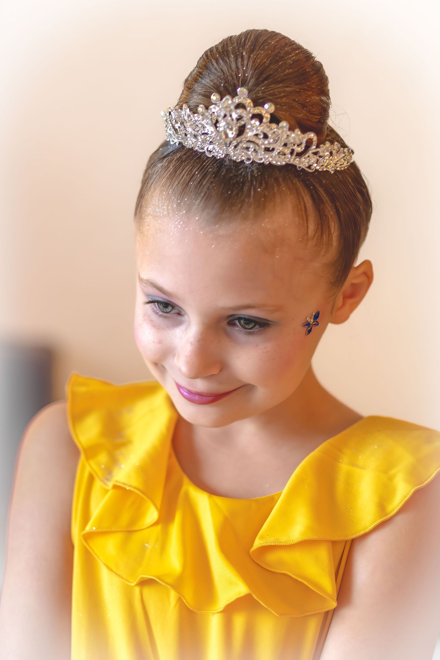 Young girl in yellow dress smiles softly wearing a sparkling tiara with a elegant ballet bun hairstyle.