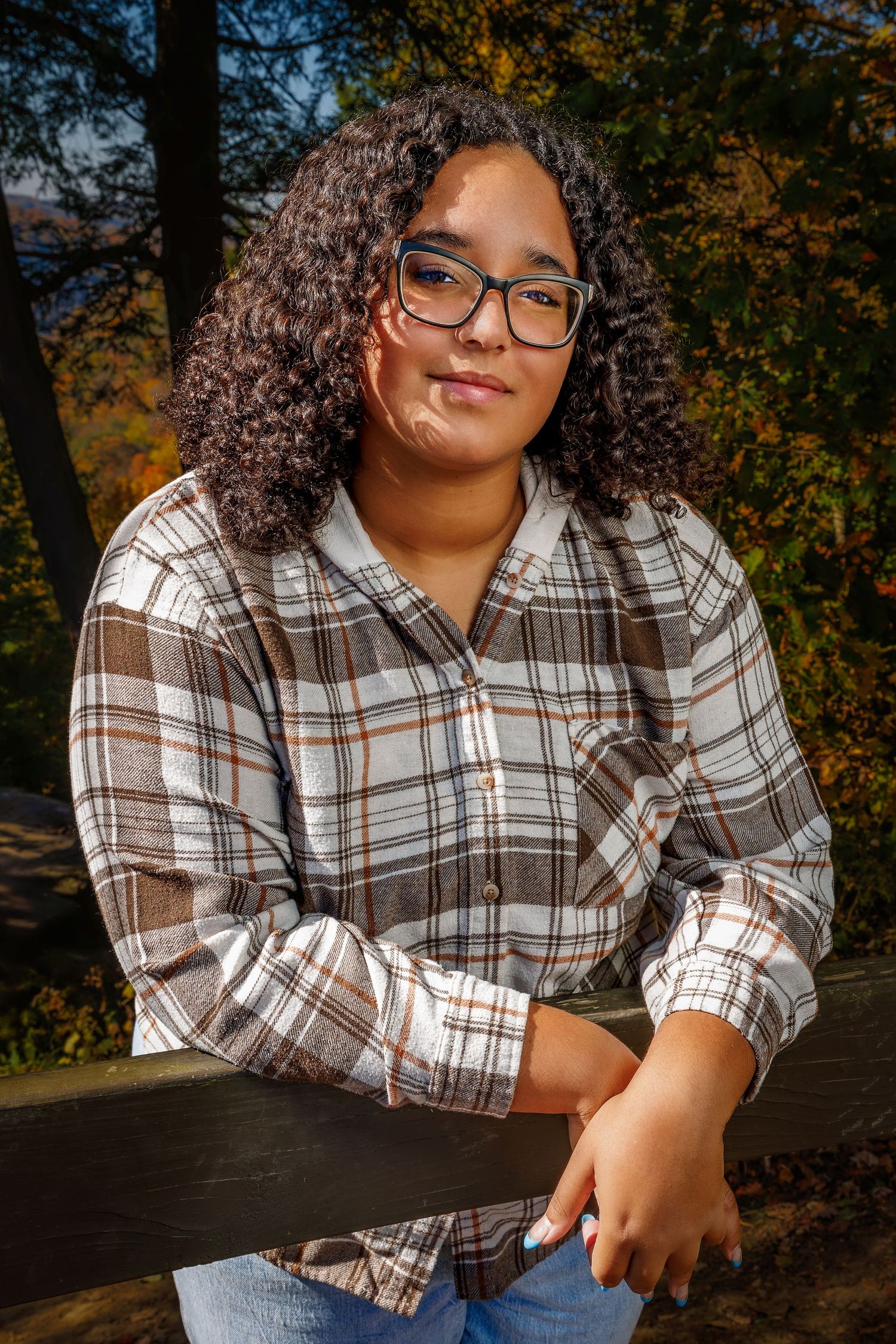 Teen girl with curly hair and glasses poses at a wooden fence surrounded by colorful autumn foliage.