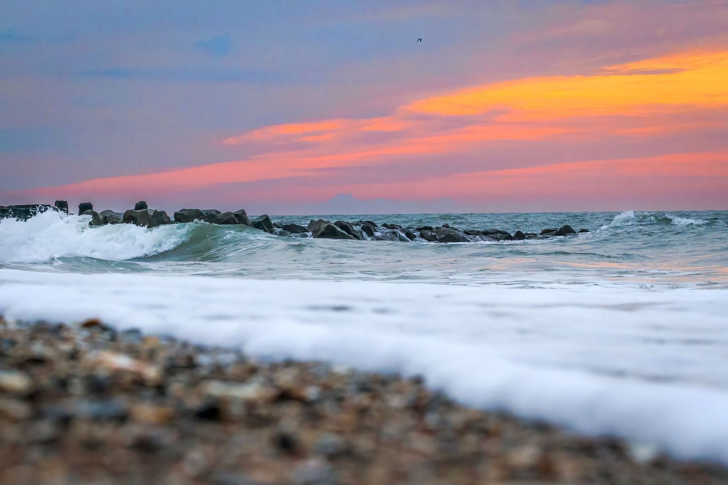 Rocky beach at sunset with crashing waves and vibrant orange and pink sky over calm ocean waters.