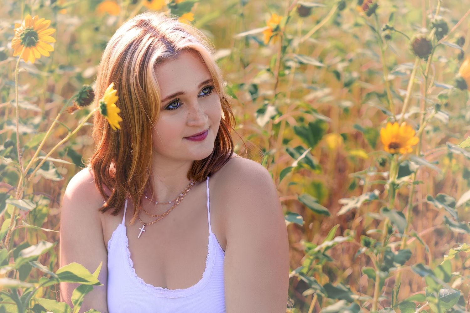 Young woman with a sunflower in her red hair poses in a sunflower field wearing a white top and cross necklace.