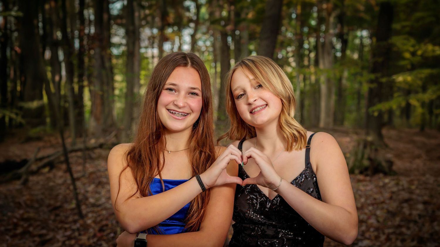 Two smiling teenage girls in formal attire make a heart shape with their hands in a forest backdrop.