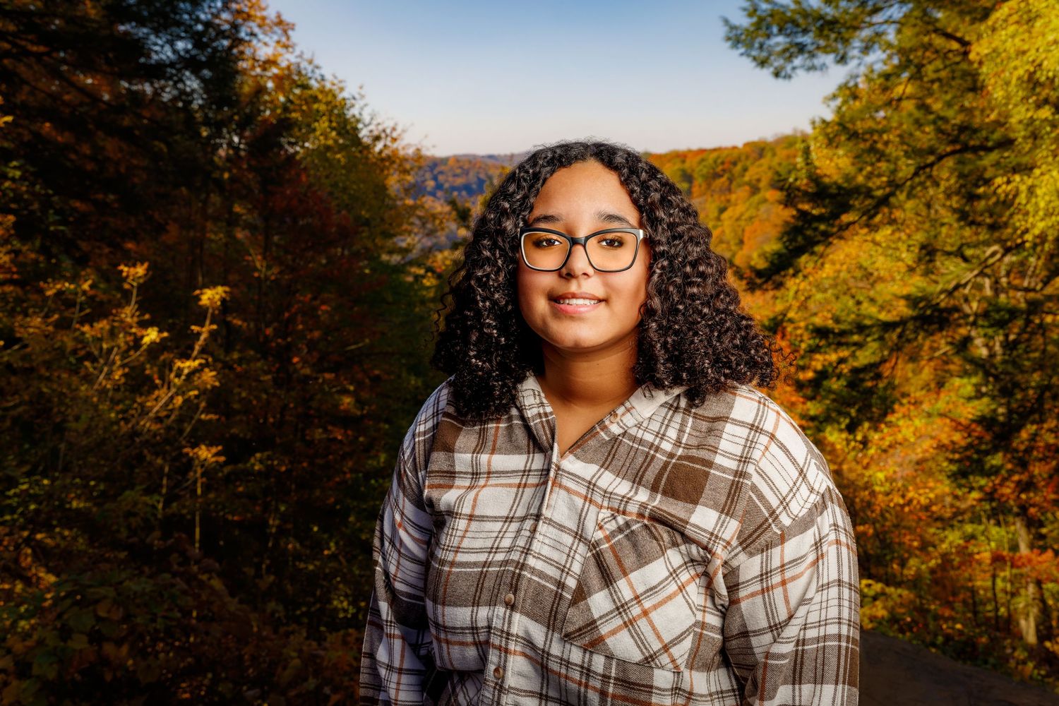 Young woman with curly hair and glasses poses outdoors among vibrant autumn foliage trees.