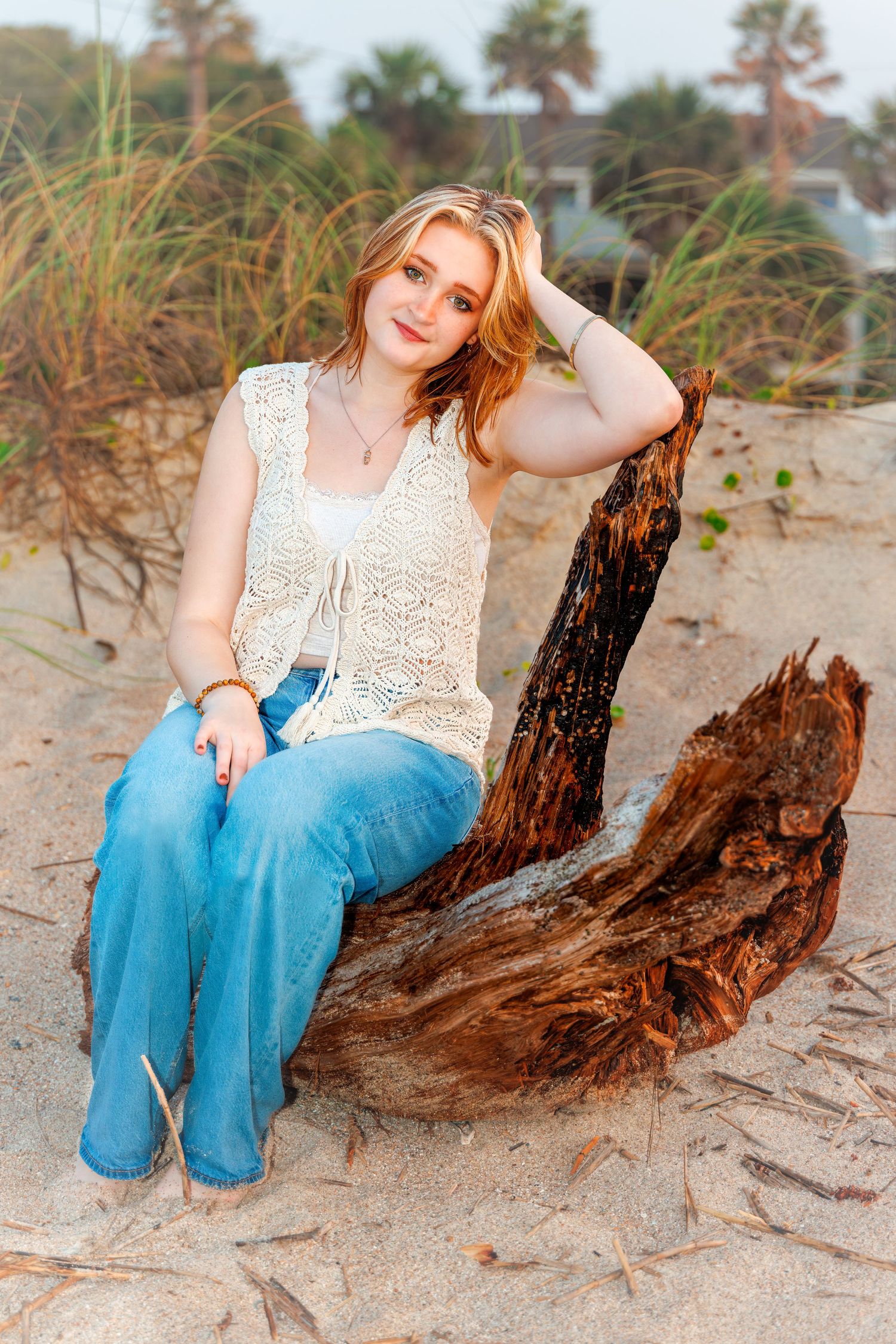 Young blonde woman in white lace top and jeans sits on driftwood during a relaxed beach portrait session.