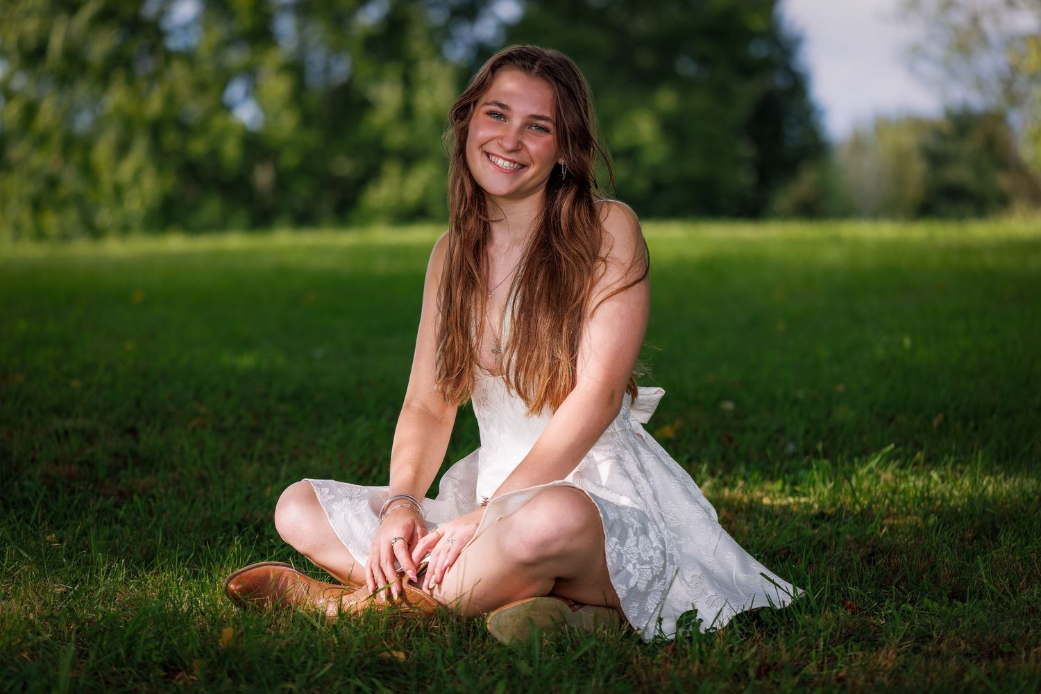 Young woman in white dress sitting cross-legged on green grass, smiling outdoors in golden sunlight.