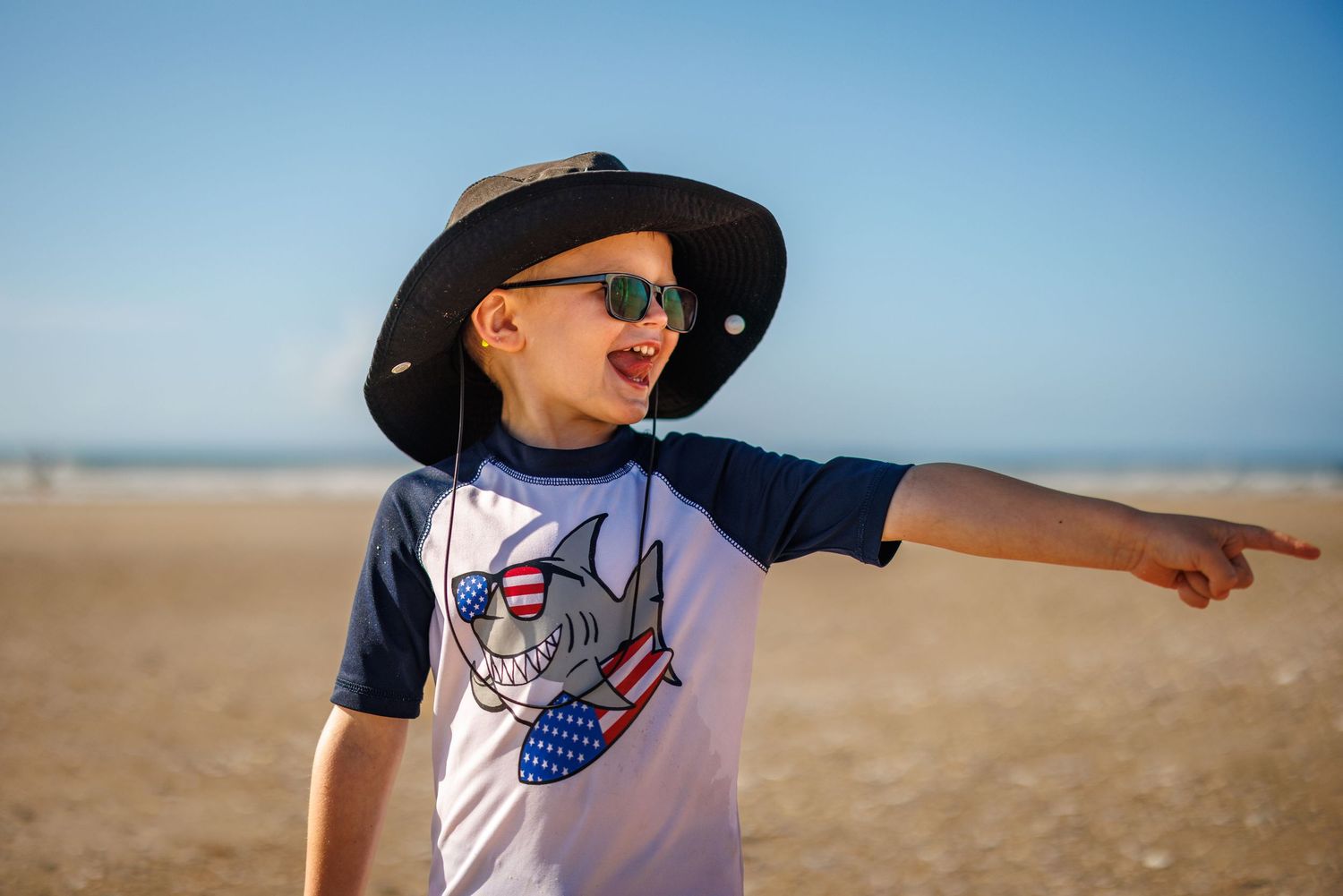 Adventurous young boy in cowboy hat and sunglasses points into the distance on a sunny beach.