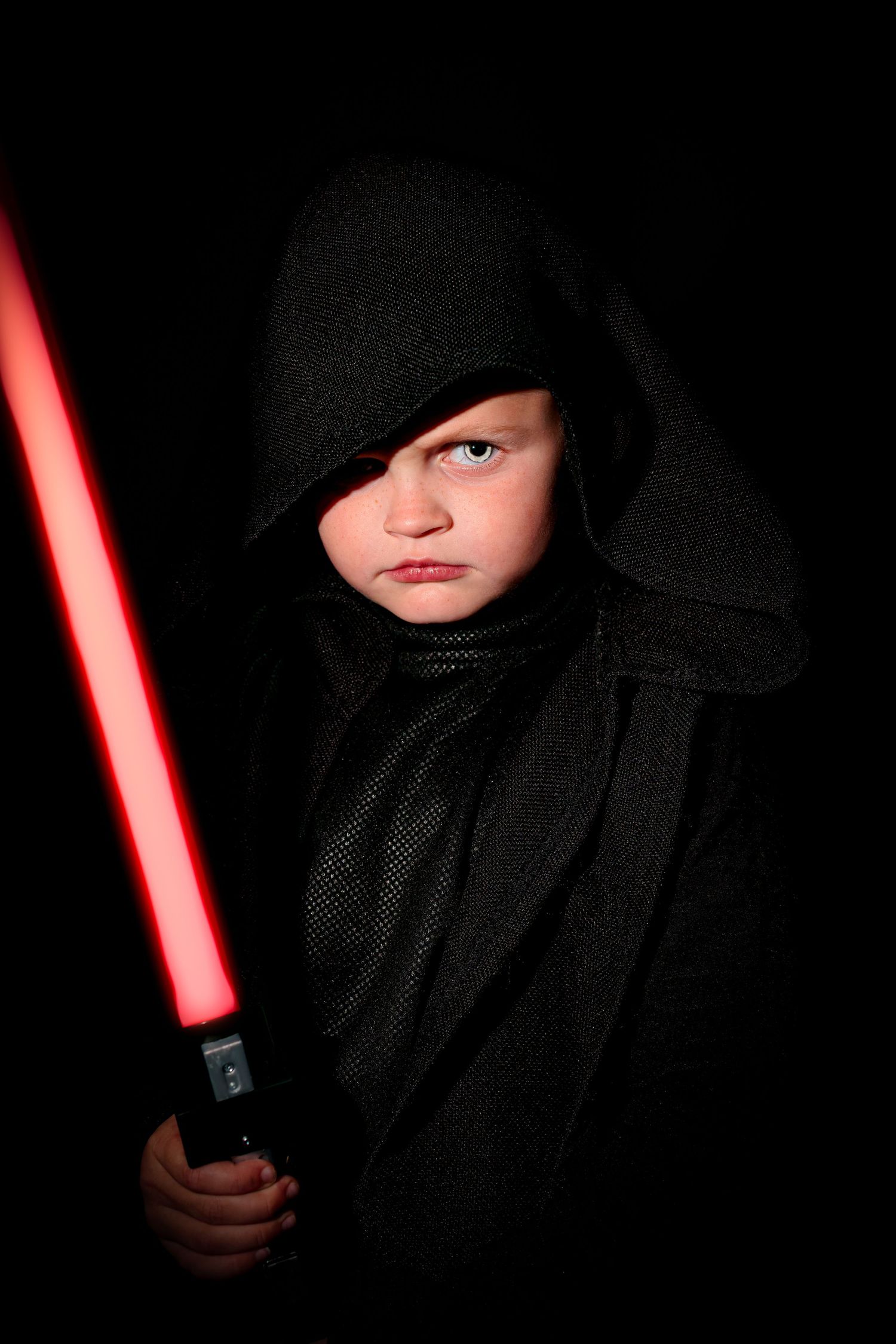 Young child dressed as a Sith lord in black hooded robe holding a red lightsaber.