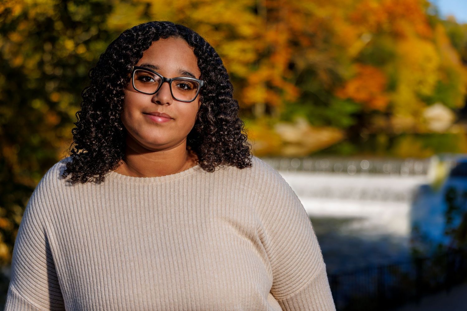 Young woman with curly hair and glasses posing outdoors with colorful autumn foliage backdrop.
