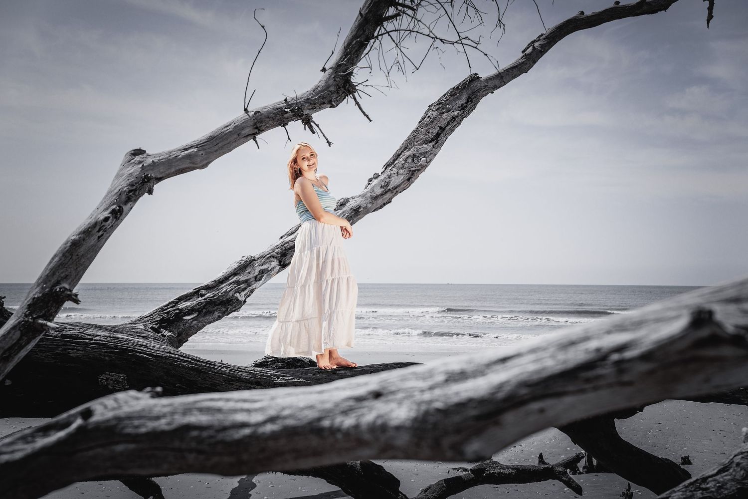 Woman in white dress posing on driftwood at the beach with ocean waves and overcast sky in background.