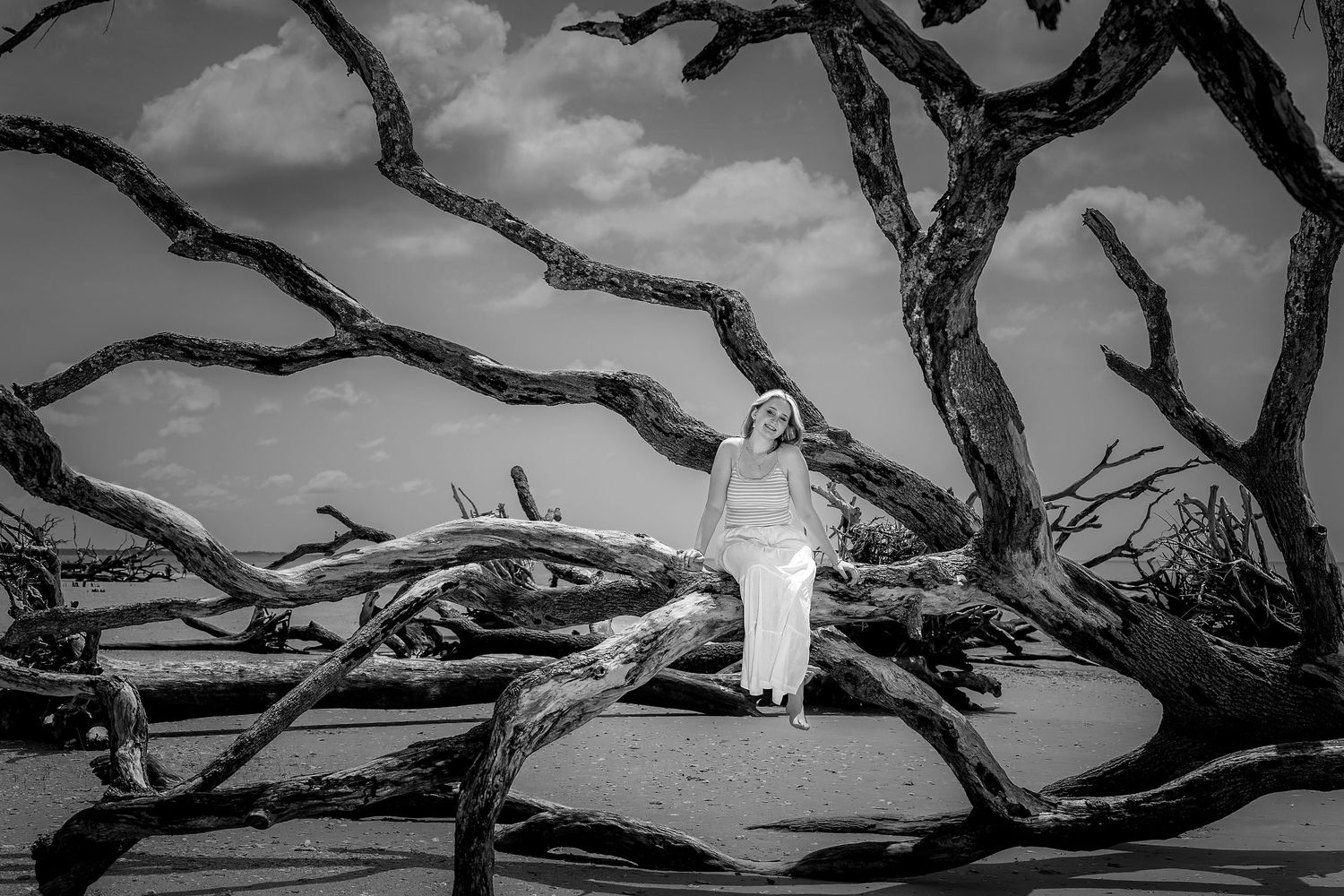 Black and white photo of a woman in white dress standing among dramatic driftwood trees on a beach.