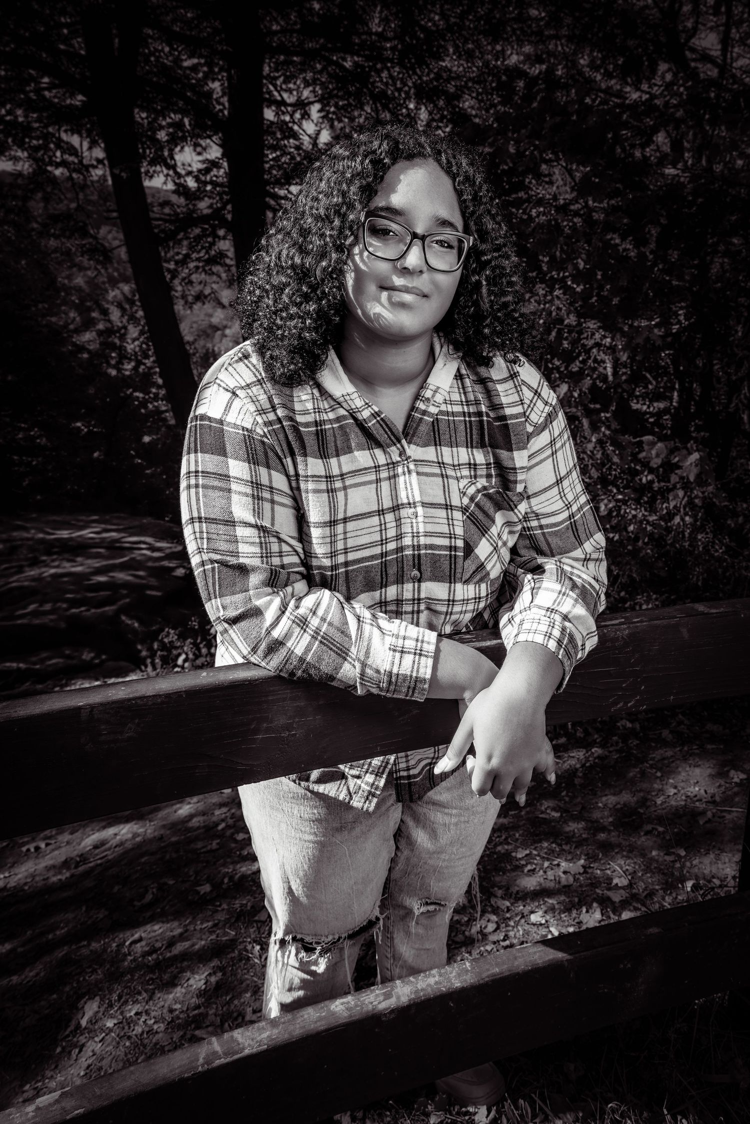 Black and white portrait of a curly-haired teen with glasses leaning on a wooden fence outdoors.