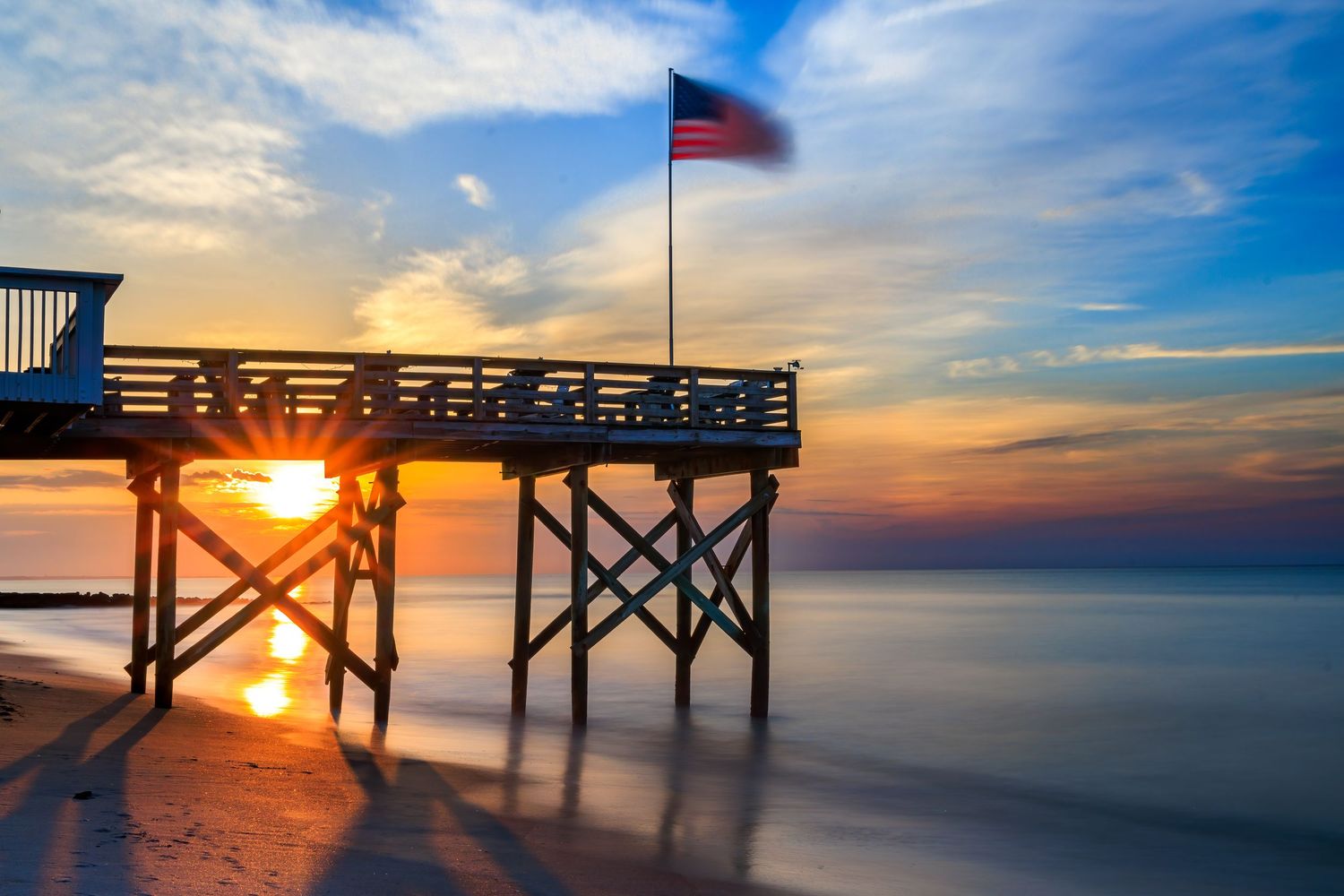 American flag waving at sunset from a wooden beach pier.