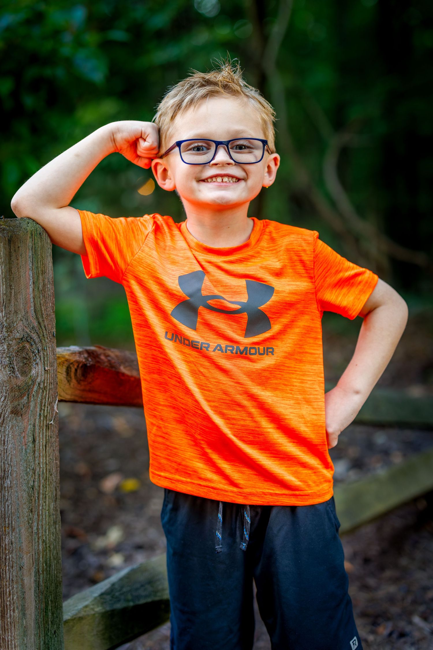 Smiling young boy in orange Under Armour shirt flexes his muscle while wearing glasses outdoors.