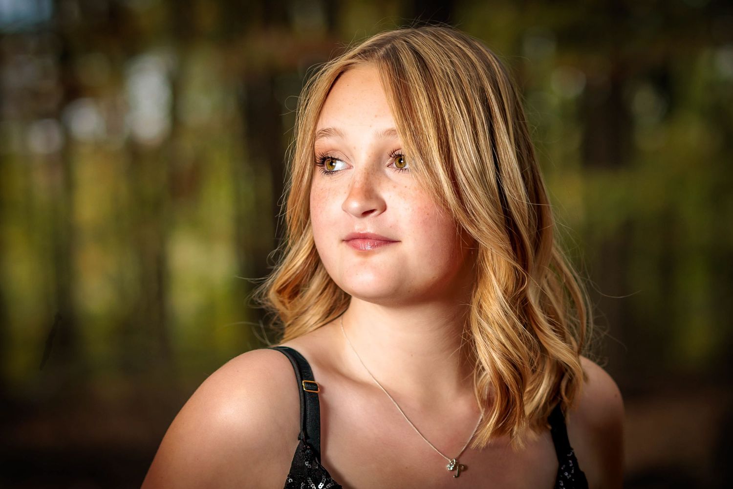 Close-up portrait of a young blonde girl with a cross necklace in a softly blurred forest setting.