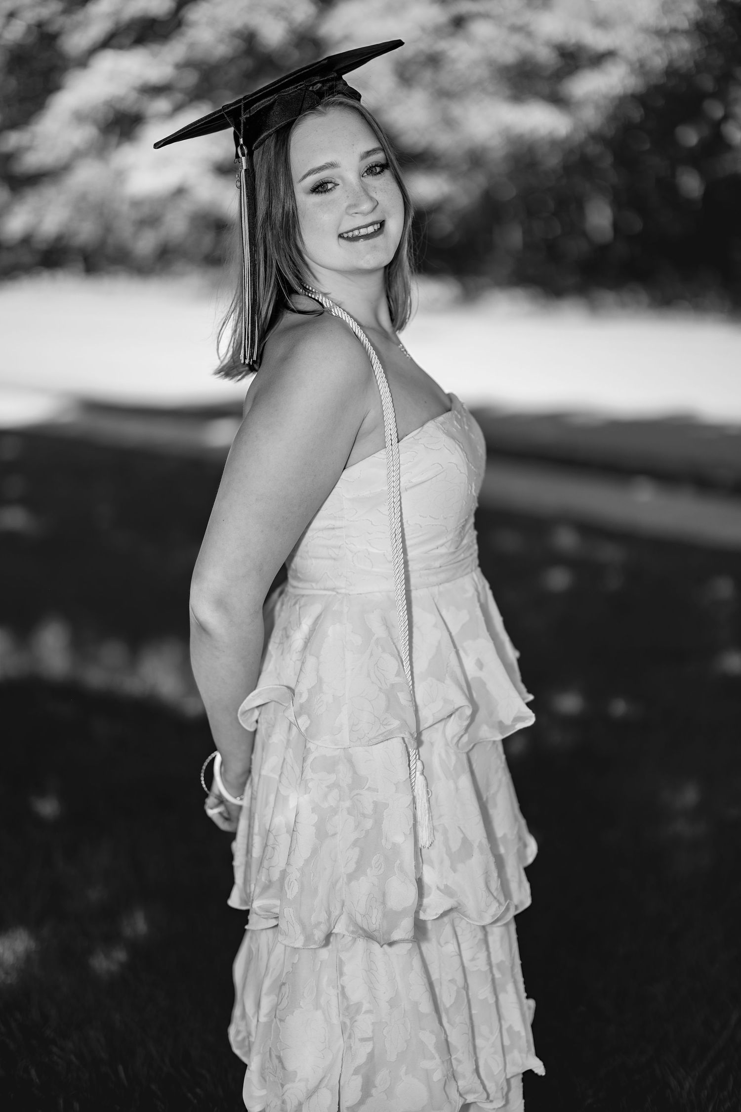 Smiling graduate in white ruffled dress and mortarboard cap poses in a classic black and white portrait.