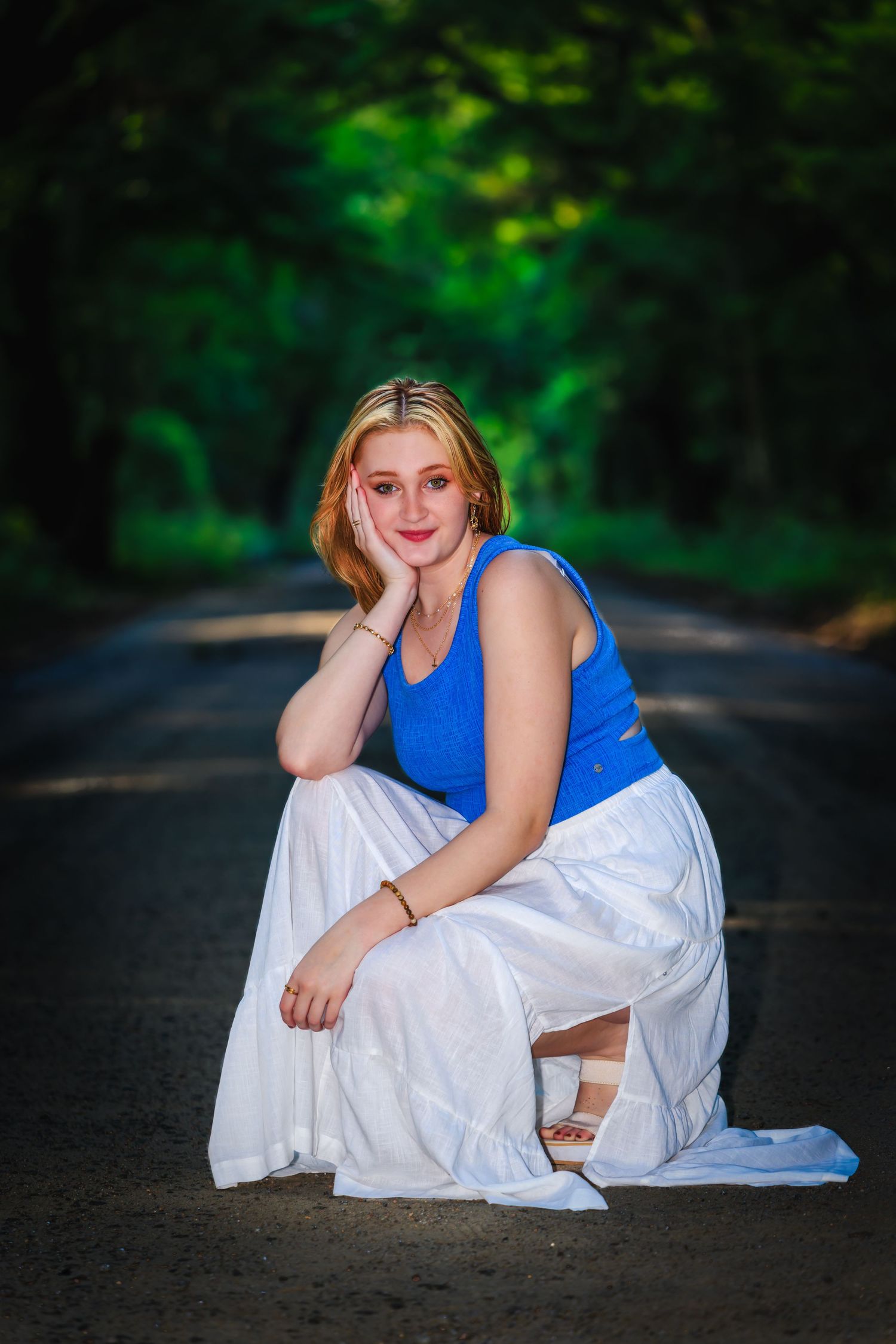 Young woman in blue tank top and white skirt poses on a tree-lined road in a serene outdoor portrait.