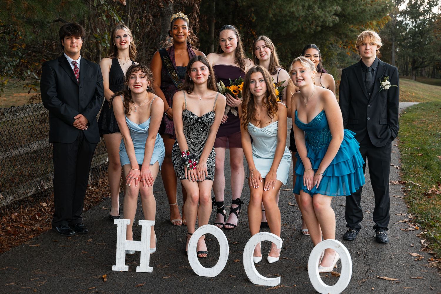 A group of smiling teenagers dressed in formal homecoming attire pose outdoors beside large HOCO letters.