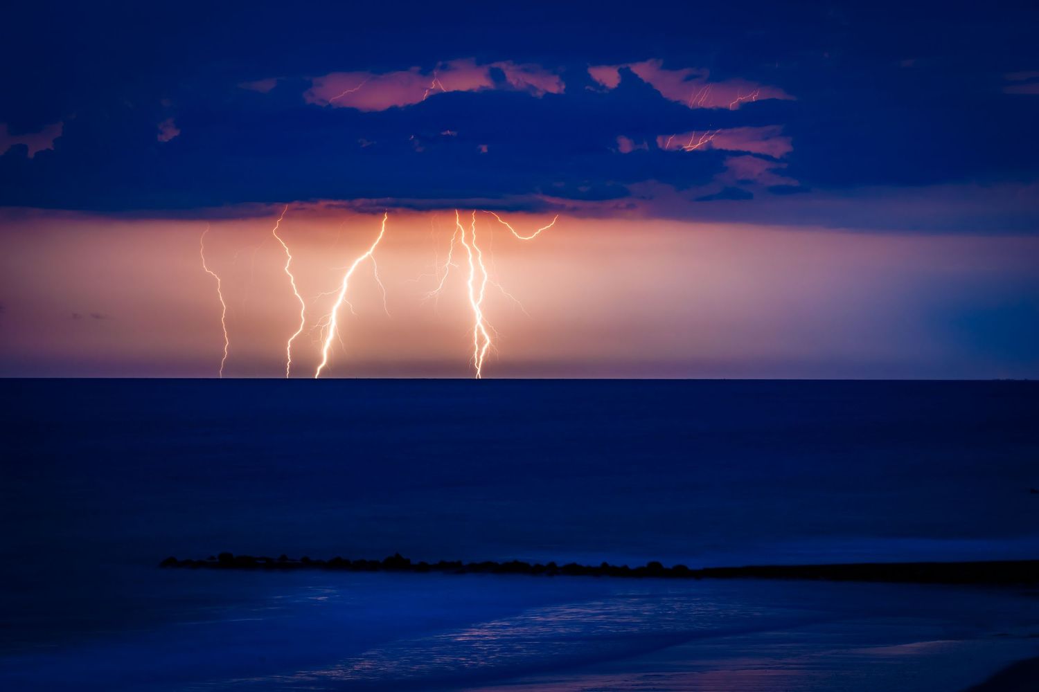 Lightning bolts strike over the ocean during a dramatic blue twilight storm.