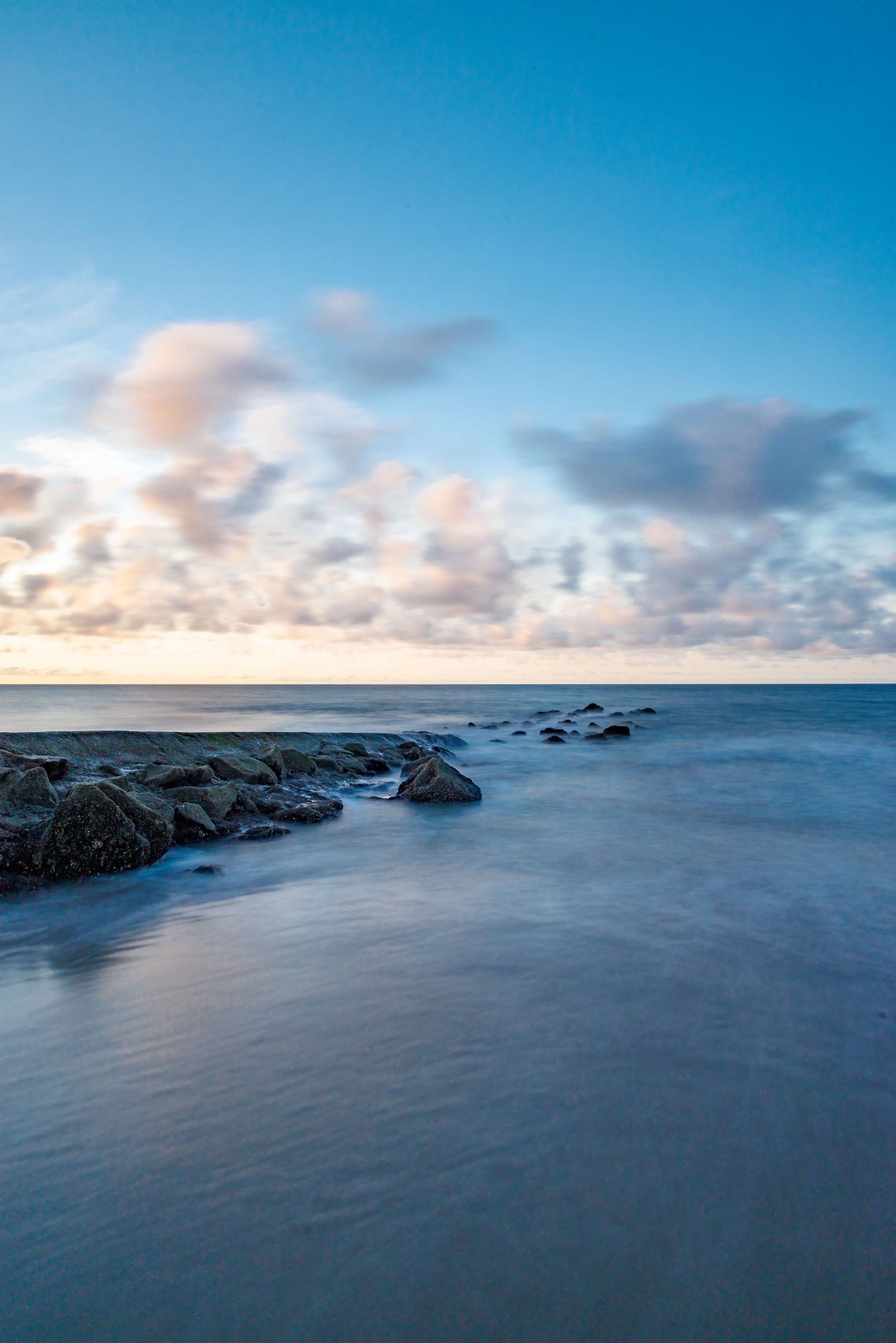 Long exposure seascape at dusk with smooth water, rocky shoreline, and dramatic blue sky with clouds.