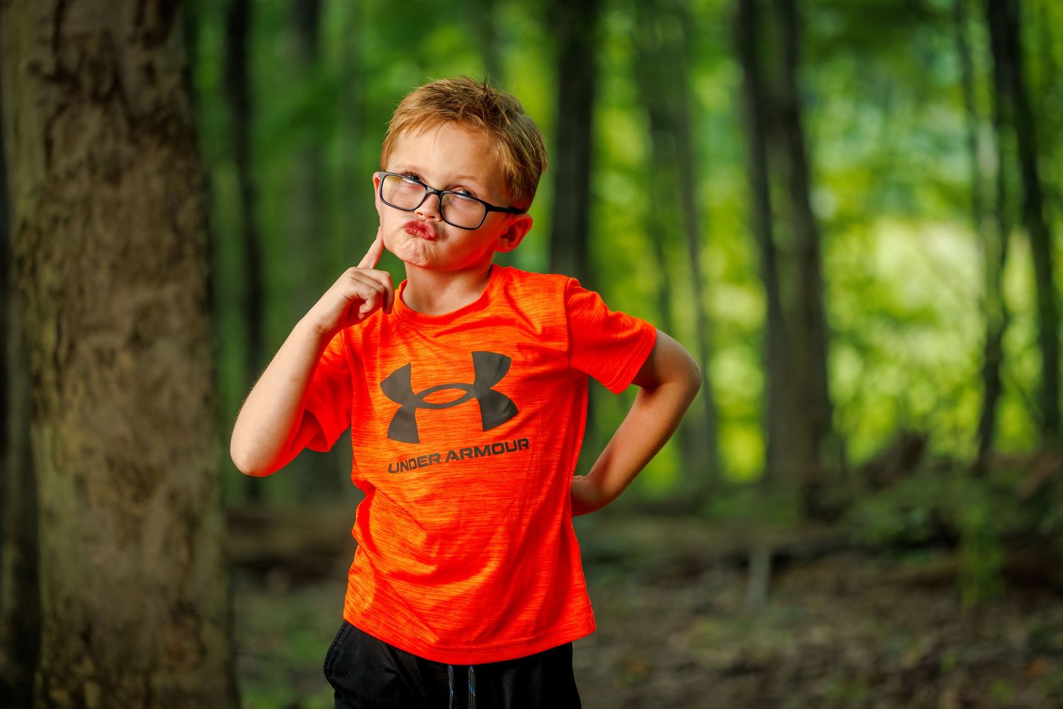 A young red-haired boy in an orange Under Armour shirt and glasses poses thoughtfully in a lush forest.