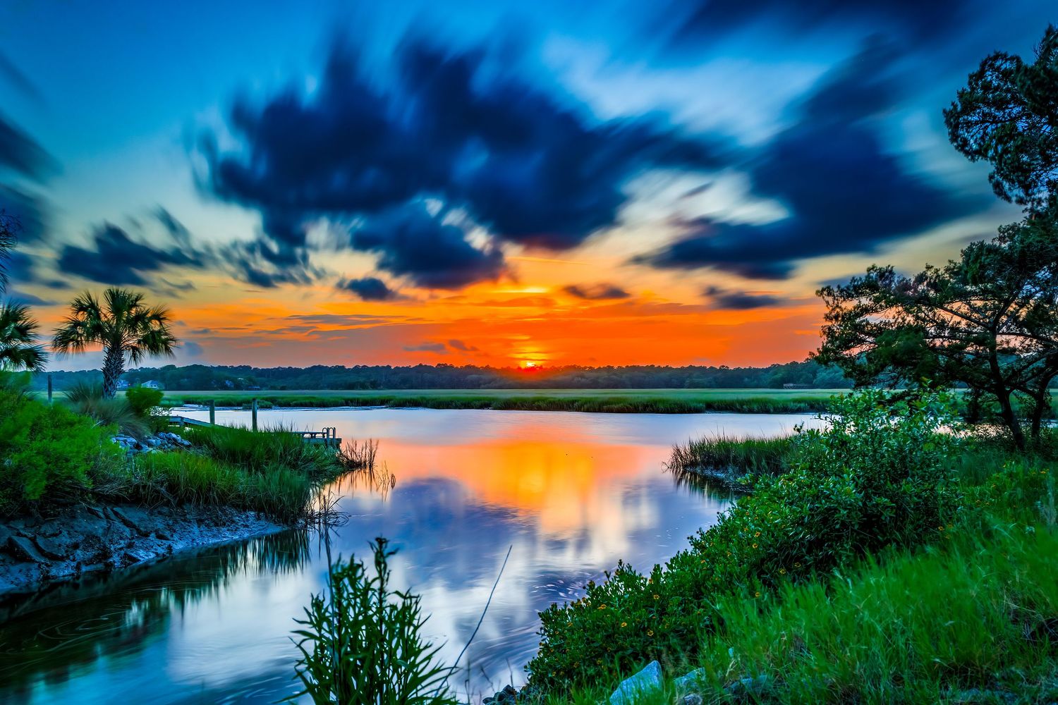 Dramatic sunset over a marsh waterway with vivid orange sky and dynamic blue storm clouds.