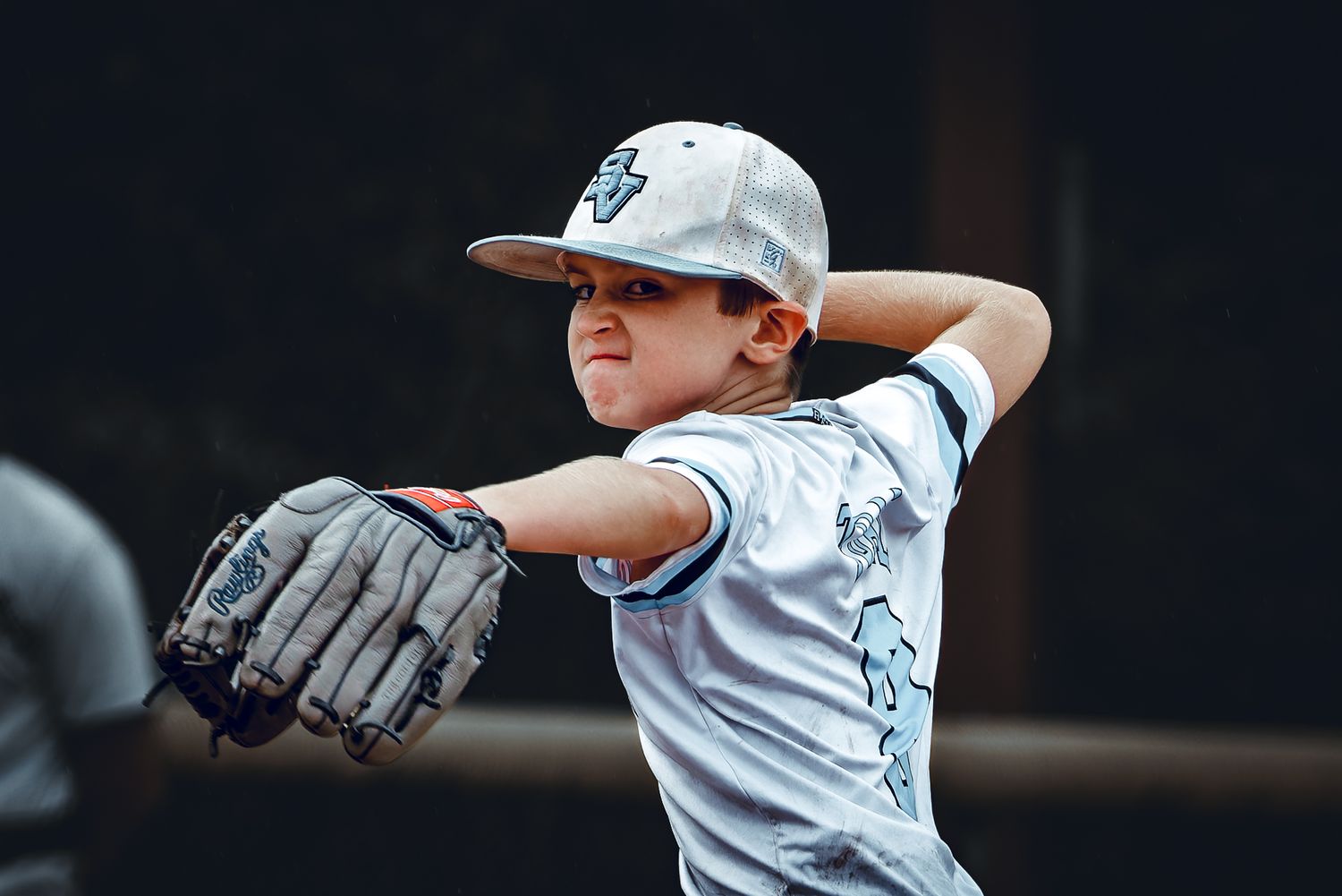 A young boy in a baseball uniform winds up to throw a pitch during a youth baseball game.